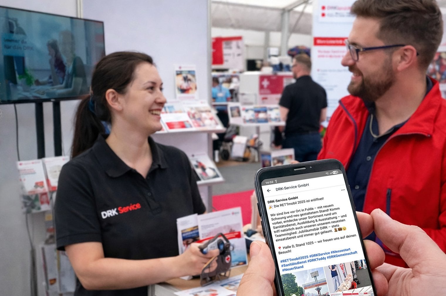 Ein Mann und eine Frau mittleren Alters stehen an einem Messestand, unterhalten sich und haben ein Lächeln im Gesicht. Im Vordergrund sieht man zwei Hände, die ein Hände halten, auf dem gerade eine Webseite zu sehen ist