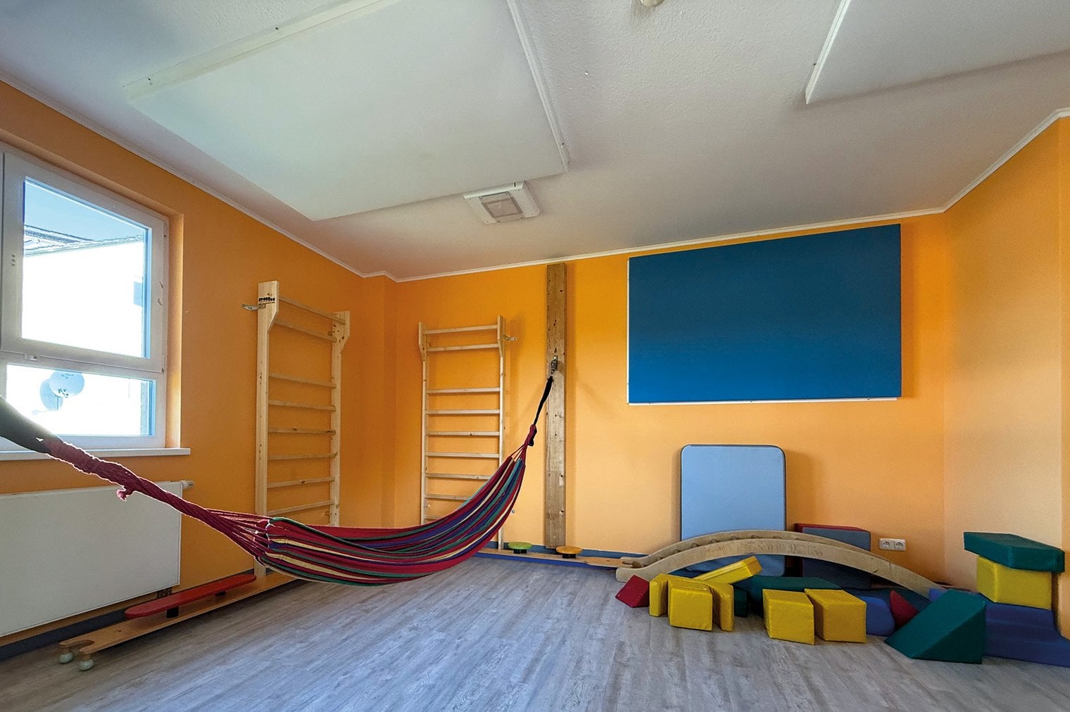 Ein Raum mit gelber Wand, Sprossenwand, Matten und einer Hängematte. An der Wand und decken hängen Paneele in Weiß und Blau.