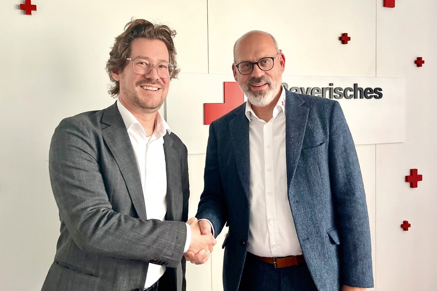 2 Männer in Sacko und weißen Hemden geben sich die Hand und lächeln in die Kamera. Im Hintergrund sieht man rote Kreuze an der Wand.