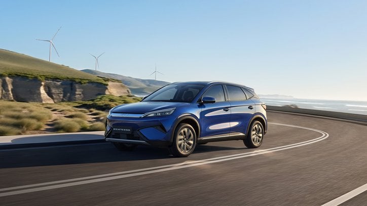 Ein blauer SUV BYD ATTO fährt auf einer kurvigen Küstenstraße entlang. Im Hintergrund sind Meer, Felsen und mehrere Windräder auf Hügeln zu sehen.