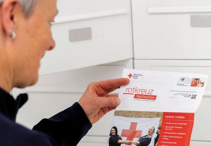 Eine Frau steht vor einem Hausbriefkasten. In den Händen hält sie einen Brief in rot-weißer Gestaltung und dem Namen rotkreuzNachrichten