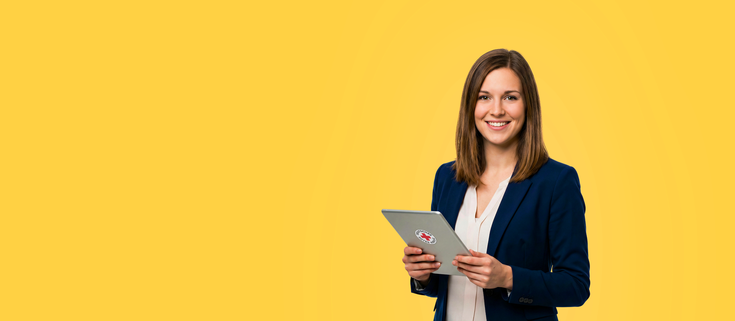 Frau in Businesskleidung hält ei Tablet in der Hand mit DRK-Rundlogo auf dem Rücken und lächelt in die Kamera.