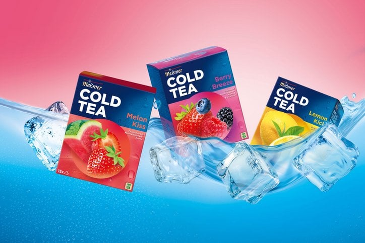 Drei Packungen Meßmer Cold Tea in den Sorten Melon Kiss, Berry Breeze und Lemon Kick schweben zwischen Eiswürfeln und spritzendem Wasser vor einem pink-blauen Hintergrund.