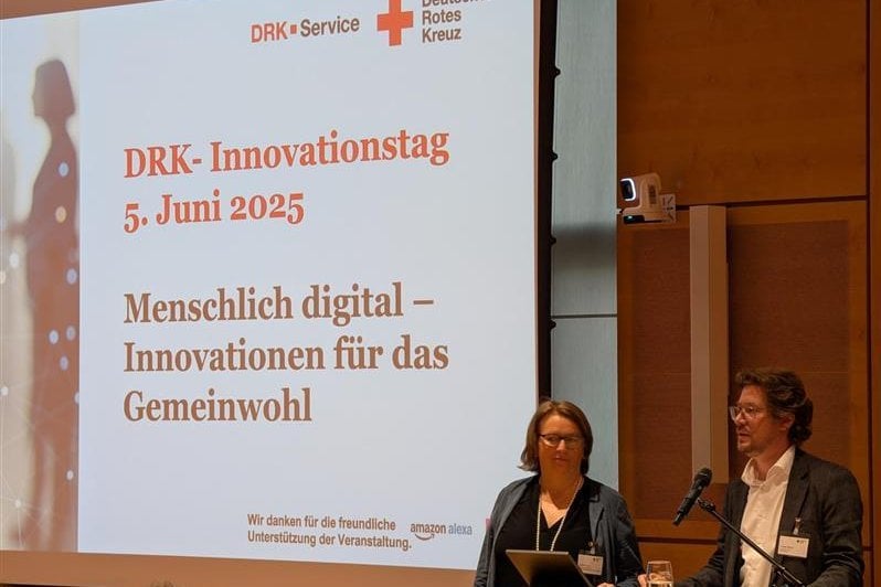 Eine Frau und ein Mann stehen an einem Rednerpult neben einer großen Leinwand mit Aufschrift "DRK-Innovationstag. 5. Juni 2025"