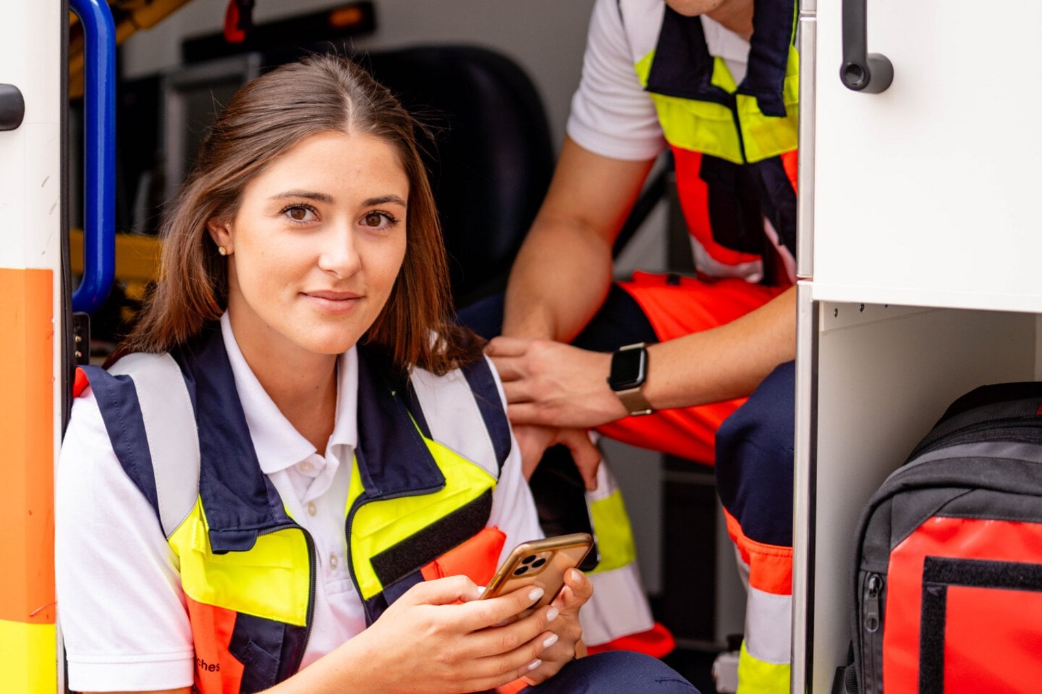 Zwei junge Rettungskräfte sitzen im Laderaum eines Rettungswagens. Die junge Frau hält ein Handy in der Hand.